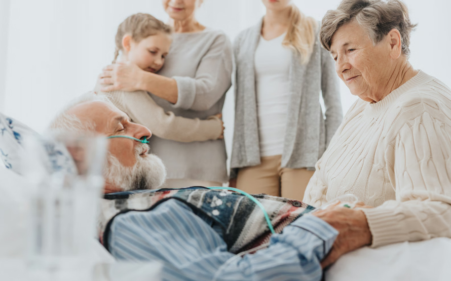 An elderly man with a white beard lies peacefully in bed with an oxygen cannula, while a senior woman holds his hand and a younger woman hugs a child in the background.