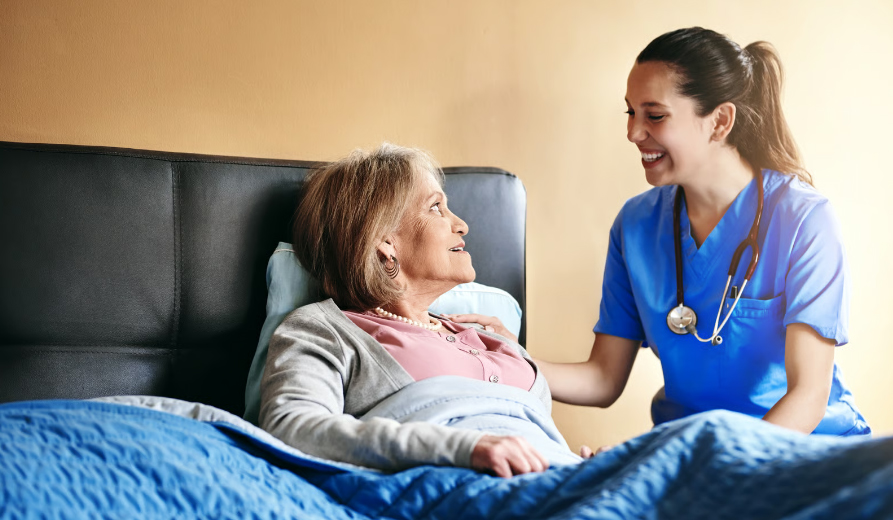 A smiling female nurse in blue scrubs sits on the edge of a bed, looking warmly at an elderly woman resting under a blue patterned blanket.