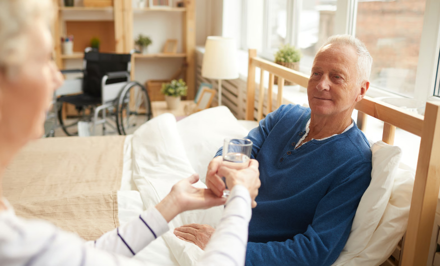 A senior man resting in a hospital bed at home receiving a glass of water from a caregiver, illustrating hospice home equipment and personalized comfort care.