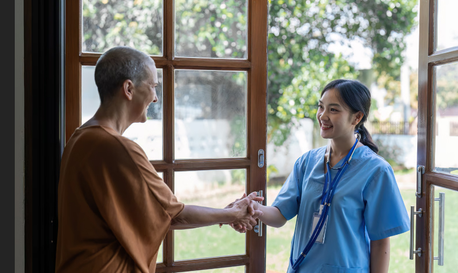A friendly hospice nurse in blue scrubs shaking hands with a smiling patient at the doorway of a home.