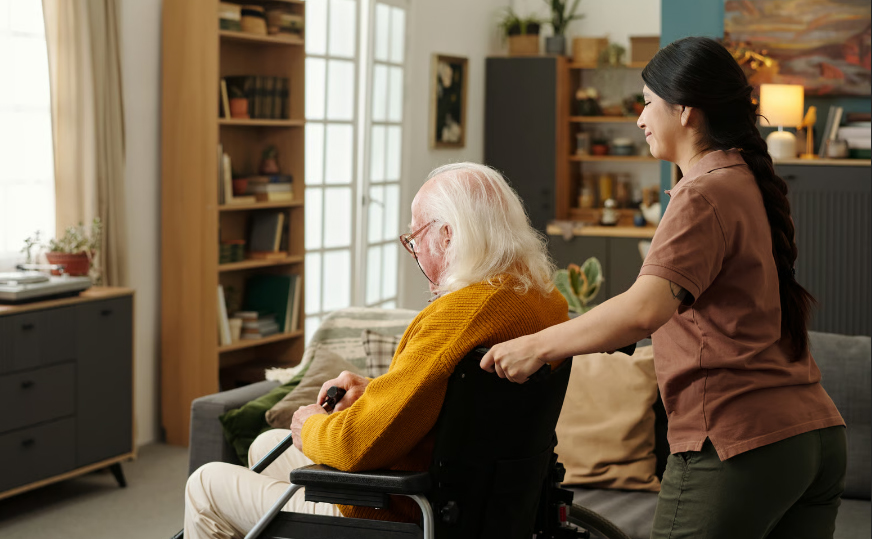 A female nurse in blue scrubs talking with a senior male veteran about hospice care options in a home setting, with a wheelchair visible in the background.