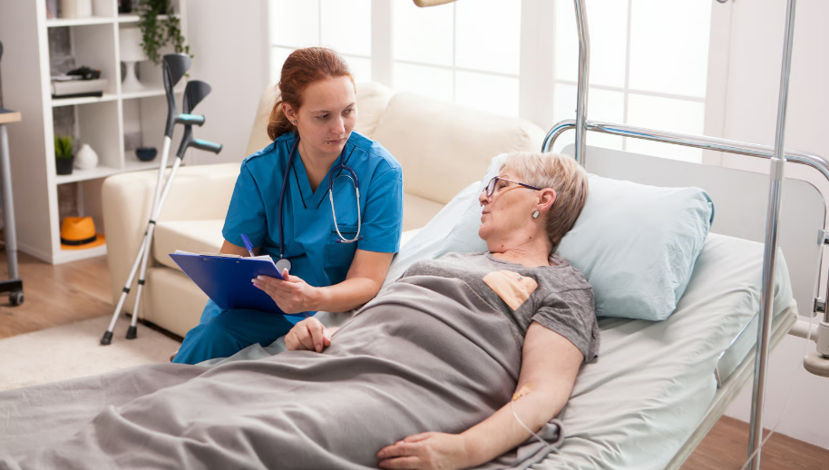 A female home nurse in blue scrubs sits beside a hospital bed at home, writing on a clipboard while speaking with an elderly woman recovering from surgery.