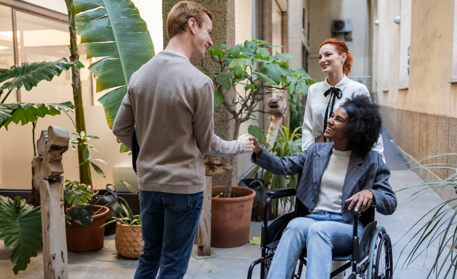 A smiling woman in a wheelchair shakes hands with a male colleague in a modern outdoor hallway filled with potted plants, while another female colleague looks on.