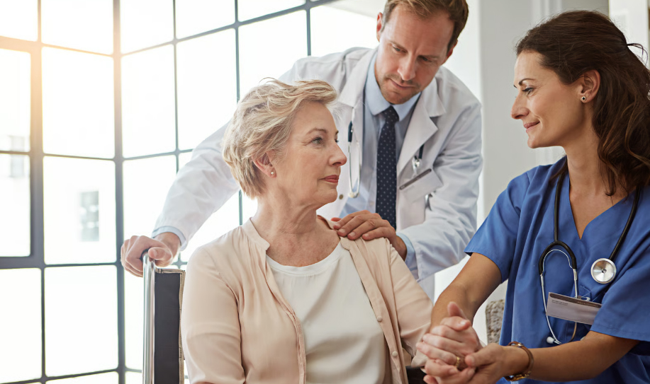 A male doctor in a white lab coat and a female nurse in blue scrubs comfort a senior woman sitting in a wheelchair near a large window.