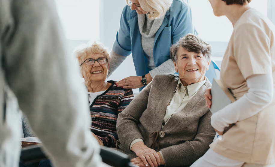 Two smiling elderly women sitting together and interacting with two supportive care facility staff members in a bright common room.