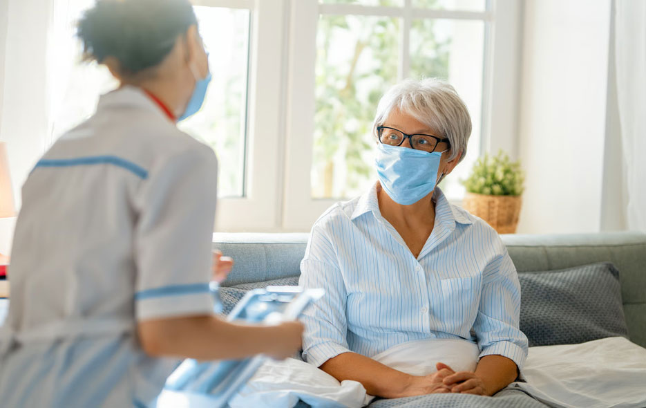 An elderly woman wearing glasses and a blue face mask sits up in bed, looking attentively at an Aspen Grove Hospice nurse who holds a clipboard, demonstrating how comfort-focused medical care is provided directly to patients in the Denver Metro area.