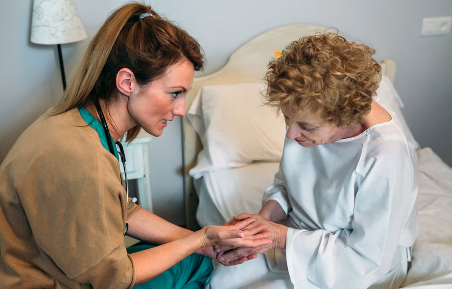 A compassionate female nurse holding the hands of an elderly woman resting in a hospital bed, illustrating the calming strategies and comfort care provided for anxiety and restlessness by Aspen Grove Hospice in the Denver Metro area.