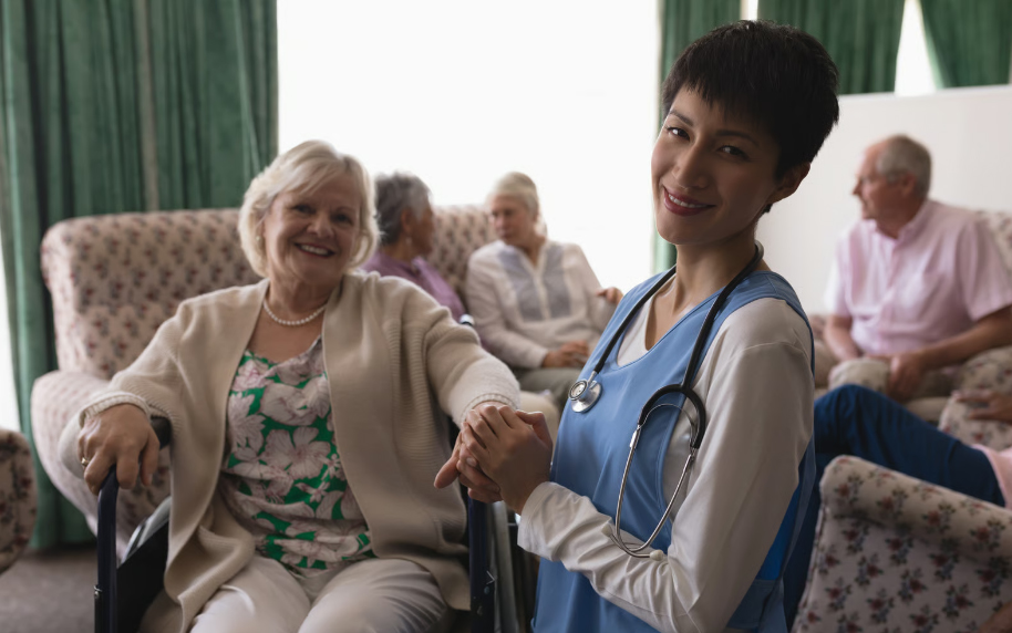 A smiling female nurse with a stethoscope holding the hand of an elderly woman in a wheelchair at a senior care facility, with other residents in the background.