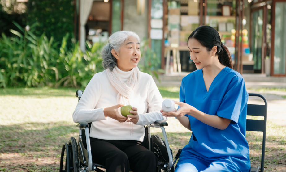 An Asian nurse in blue scrubs explaining prescription bottle labels to an elderly woman in a wheelchair outdoors.