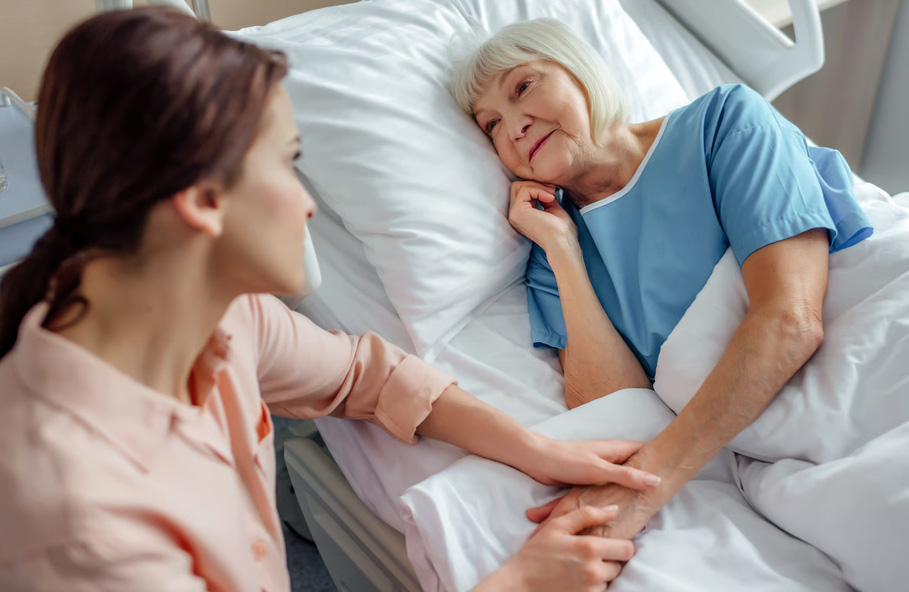 A compassionate caregiver holding the hand of an elderly woman lying in a hospital bed.
