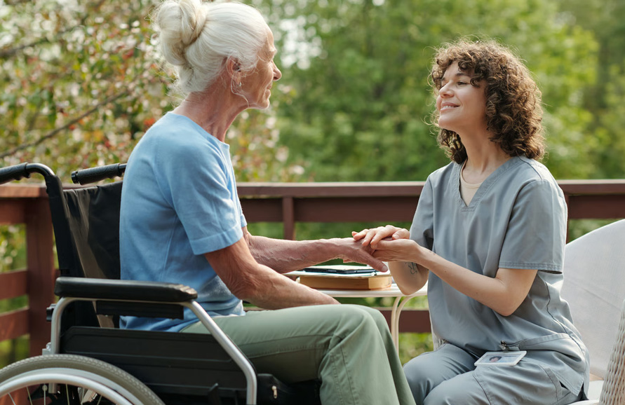 A compassionate hospice caregiver in Denver holding the hand of a smiling elderly patient in a wheelchair, illustrating emotional and physical comfort.