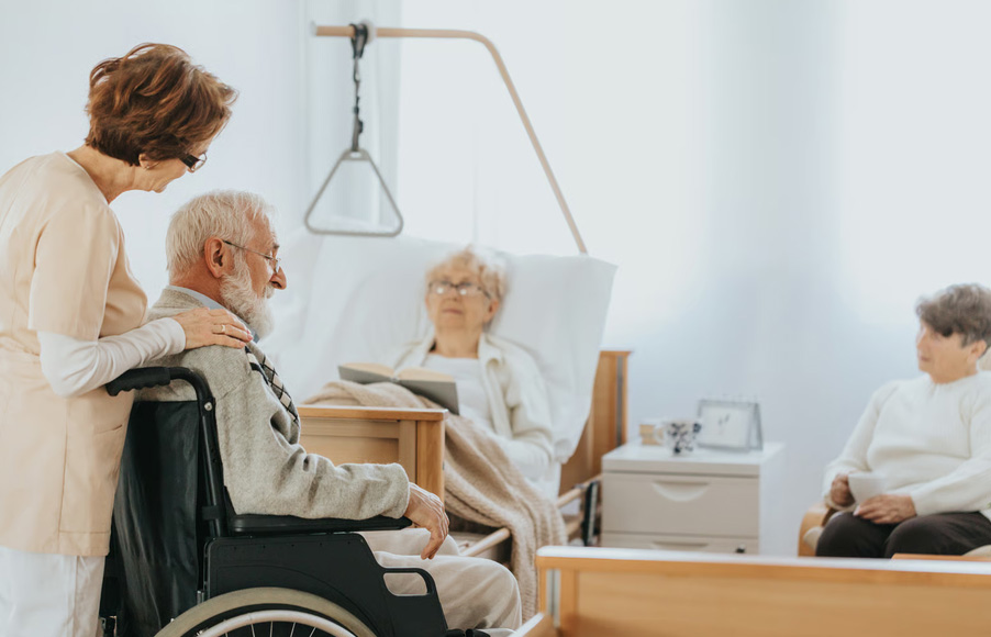 A compassionate caregiver placing a hand on the shoulder of an elderly man in a wheelchair in a room with a medical hospital bed.