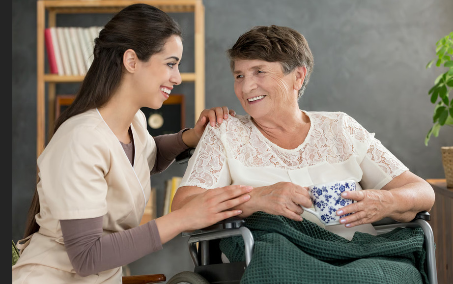 A smiling hospice caregiver placing a comforting hand on an elderly woman sitting upright in a wheelchair, illustrating non-pharmacological breathlessness management.