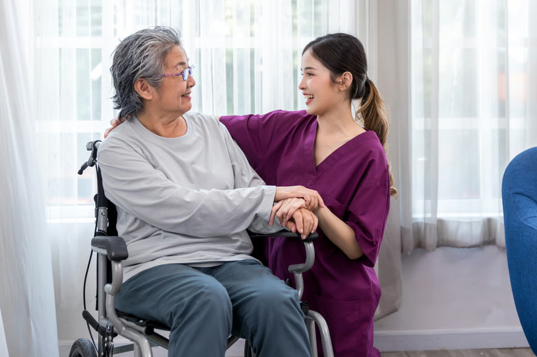 A cheerful hospice caregiver in purple scrubs kneeling next to a smiling senior woman in a wheelchair, holding her hands.