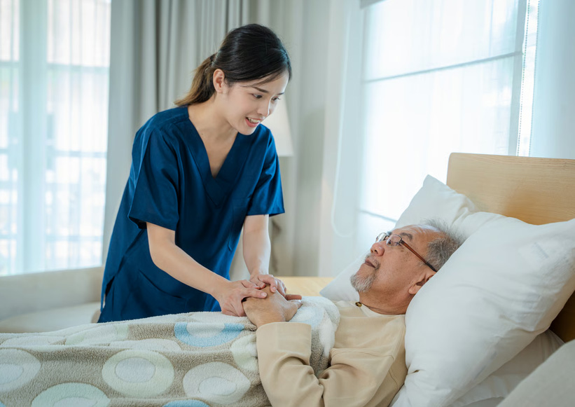 A caring hospice nurse in blue scrubs holding the hand of an elderly patient lying in a home hospital bed.